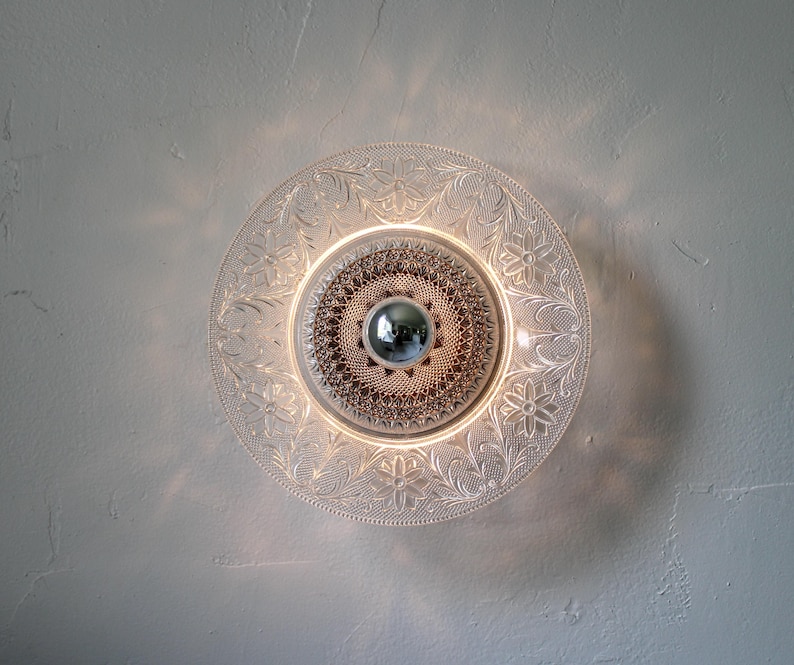 May include: A clear glass wall sconce with a decorative floral and leaf pattern. The light fixture has a central, textured design and a silver-colored, spherical bulb. The sconce is mounted on a light gray wall.