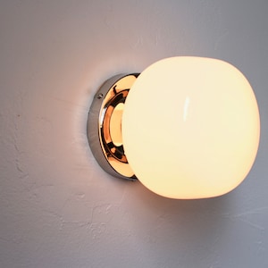 Op de afbeelding: Een chromen wandlamp met een witte glazen bolvormige kap. Het licht is aan en geeft een warm licht.