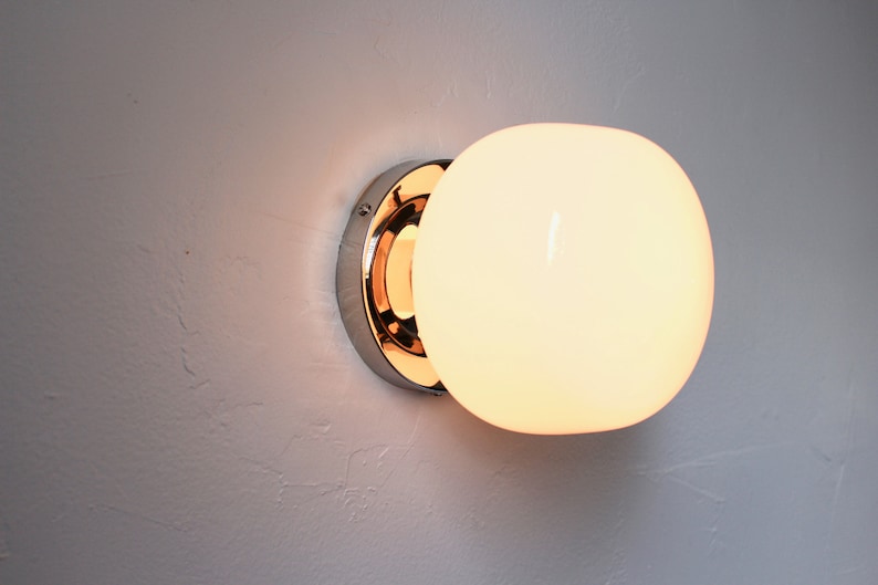Op de afbeelding: Een chromen wandlamp met een witte glazen bolvormige kap. Het licht is aan en geeft een warm licht.