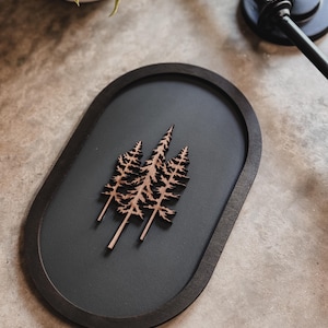 May include: An oval, dark gray wooden tray with a raised, dark brown border. Three stylized, light brown wooden pine trees are centered on the tray. The tray is on a neutral-toned surface.