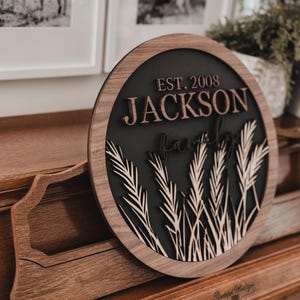 以下が含まれることがあります： 「EST. 2008 JACKSON Family」の文字が重ねてデザインされた丸い木製の看板。看板は、麦の穂と明るい木製の縁取りのある暗い背景が特徴です。看板は木製の表面に表示されています。