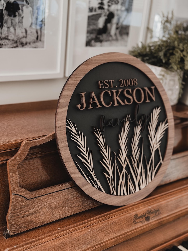 以下が含まれることがあります： 「EST. 2008 JACKSON Family」の文字が重ねてデザインされた丸い木製の看板。看板は、麦の穂と明るい木製の縁取りのある暗い背景が特徴です。看板は木製の表面に表示されています。
