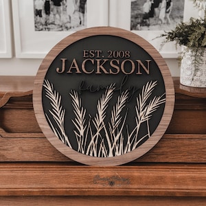 以下が含まれることがあります： 「JACKSON」と「EST. 2008」の文字が重ねてデザインされた、丸い木製のファミリーサイン。濃い色の背景に麦のようなアクセントと天然木のフレームが特徴です。木製のピアノの上に飾られています。