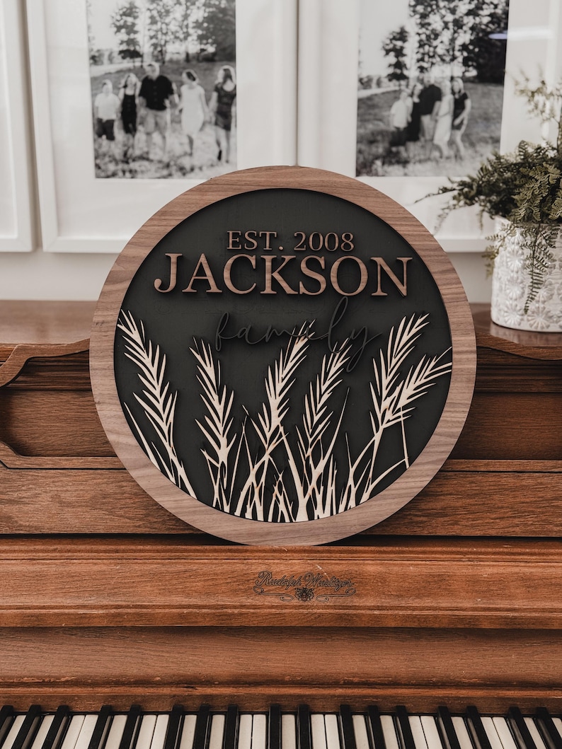 以下が含まれることがあります： 「JACKSON」と「EST. 2008」の文字が重ねてデザインされた、丸い木製のファミリーサイン。濃い色の背景に麦のようなアクセントと天然木のフレームが特徴です。木製のピアノの上に飾られています。