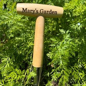 May include: A garden tool with a wooden handle and metal tip, with the text "Mary's Garden" engraved on the handle. The tool is set amongst green foliage. The handle is light brown in colour.