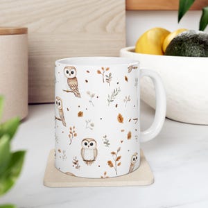 Könnte beinhalten: Weiße Keramiktasse mit einem sich wiederholenden Muster aus Aquarell-Eulen, Blättern und Zweigen in Braun- und Beigetönen. Die Tasse hat einen gebogenen Henkel und steht auf einem beigen Untersetzer. Die Tasse ist für Heißgetränke.