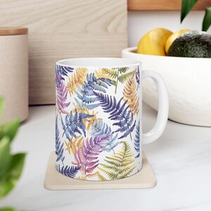 May include: White ceramic mug with a colorful fern pattern. The ferns are painted in shades of blue, purple, yellow, and green. The mug has a white handle.