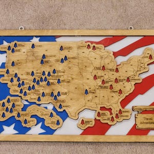May include: A rectangular wooden wall map of the United States featuring a red, white, and blue flag background. The map includes laser-cut wooden state outlines with blue and red tree icons marking locations and a sign labeled Travel Adventures.