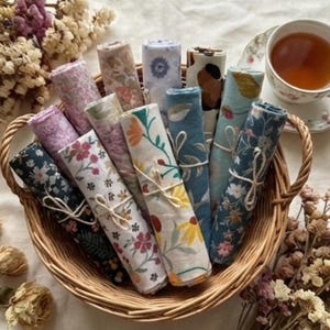 Puede incluir: Una cesta de mimbre llena de servilletas de tela enrolladas con varios estampados florales y colores, atadas con hilo. Las servilletas incluyen diseños con flores, hojas y estampados abstractos. Una taza de té y un platillo con té están en el fondo.