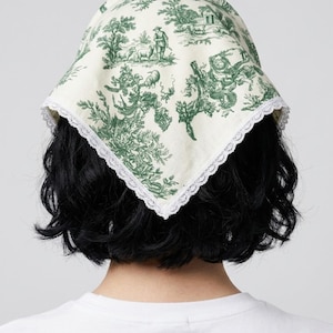May include: A triangular headscarf featuring a cream-colored fabric with a green toile de jouy print depicting pastoral scenes, including figures and trees. The edges of the headscarf are finished with a delicate white lace trim border.