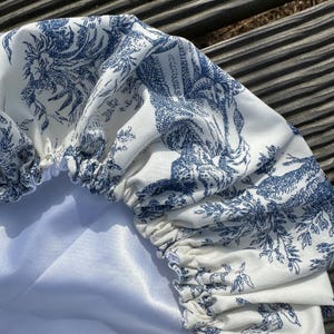 May include: A white and blue toile fabric shower cap. The cap features a repeating pattern of classical figures and foliage in a blue hue. The cap has an elastic band and a white satin lining.