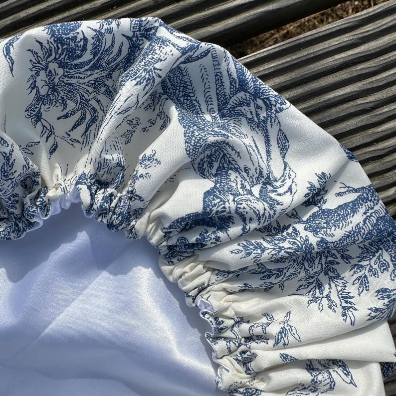 May include: A white and blue toile fabric shower cap. The cap features a repeating pattern of classical figures and foliage in a blue hue. The cap has an elastic band and a white satin lining.