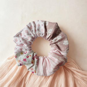 Puede incluir: Una goma para el pelo floral con una mezcla de estampados y colores. La goma presenta secciones de estampados florales rosas y blancos, un estampado a rayas rosa claro y un diseño floral malva. La tela está fruncida, creando una forma circular voluminosa.