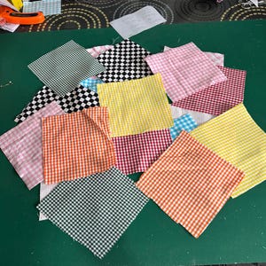 May include: A selection of gingham fabric squares in shades of orange, pink, yellow, green, and black and white. The squares are arranged on a green surface, displaying a variety of patterns and colours.