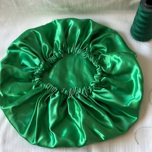 Op de afbeelding: Een felgroene satijnen slaapmuts. De muts is bovenaan gerimpeld en heeft een gladde, glanzende afwerking.
