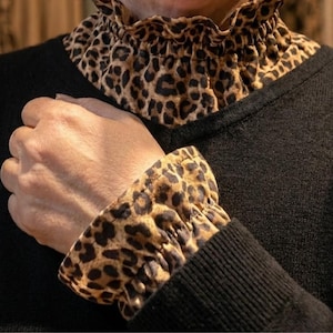 Pode incluir: Camisola preta com gola e punhos com estampa de leopardo. A gola e os punhos são feitos de um tecido com estampa de animal marrom e preto. A camisola tem um design simples e clássico.