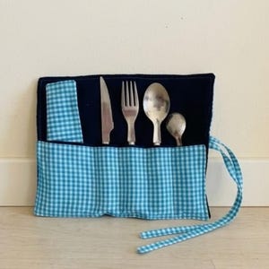 May include: A navy blue fabric cutlery roll featuring a light blue and white gingham pattern on the exterior pocket and a matching napkin. The roll contains a stainless steel knife, a fork, a large spoon, and a small spoon tucked into individual slots.