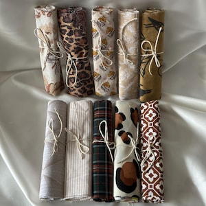 Puede incluir: Una colección de diez piezas de tela enrolladas y atadas con hilo. Las telas muestran varios patrones, incluyendo diseños florales, de leopardo, a cuadros y geométricos. La paleta de colores incluye tonos beige, marrón y verde.