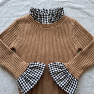 Può includere: Un maglione a coste color cammello con collo alto a balze e polsini svasati. Il colletto e i polsini presentano un motivo a quadretti bianchi e neri. Il maglione ha maniche lunghe e una silhouette aderente.