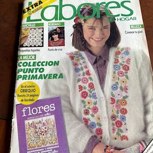 May include: A vintage Labores del Hogar magazine from March 1989, featuring a white knit cardigan with colorful floral embroidery on the front. The cover displays text including Labores del Hogar, Extra 100 Modelos, and a purple inset titled flores bordadas con DMC.