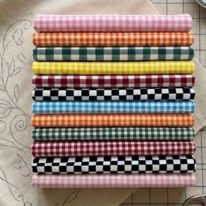 May include: A stack of rolled fabric in various colors, each with a gingham pattern. Colors include pink, orange, green, yellow, red, black and white, blue, and burgundy. The fabric is neatly stacked on a neutral-colored surface.