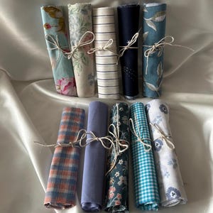 Op de afbeelding: Een verzameling opgerolde stoffen bundels, vastgebonden met touw. De stoffen hebben verschillende patronen, waaronder bloemen, strepen, ruiten en effen kleuren. Het kleurenpalet omvat blauw, roze en neutrale tinten, wat een vintage esthetiek creëert.