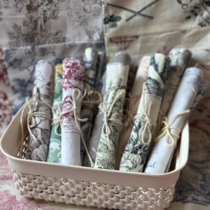 Può includere: Una collezione di tovaglioli di stoffa arrotolati in un cestino color crema. I tovaglioli presentano vari motivi, tra cui disegni floreali e paesaggistici, e sono legati con spago. Il cestino è di colore rosa chiaro e ha un design decorativo a trama aperta.