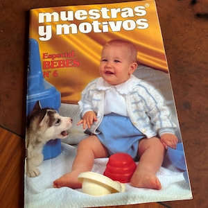 May include: A magazine cover titled Muestras y motivos Especial BEBES N 6 featuring a baby wearing a white collared shirt, a light blue and white knit cardigan, and blue shorts. A small husky puppy and red and cream plastic toys are visible.