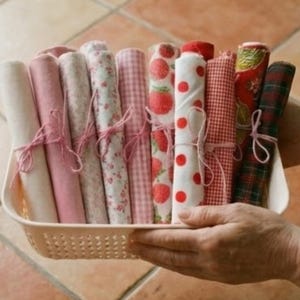 May include: A basket holds several rolls of fabric in various patterns and colors, including pink, white, and red. Some rolls feature floral, strawberry, polka dot, and plaid designs. Each roll is tied with pink twine.