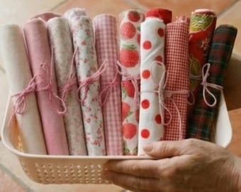 May include: A basket holds several rolls of fabric in various patterns and colors, including pink, white, and red. Some rolls feature floral, strawberry, polka dot, and plaid designs. Each roll is tied with pink twine.