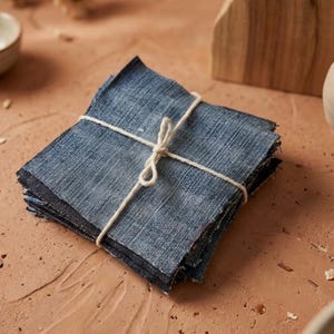 May include: A stack of square denim fabric pieces tied with a natural twine bow. The denim is a medium blue color with visible texture. The pieces are neatly stacked, suggesting a craft or sewing project.