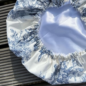 May include: A white and blue toile-patterned bonnet. The bonnet features a blue pastoral scene print on a white background. The interior is a solid white satin material. The edges are gathered with elastic.