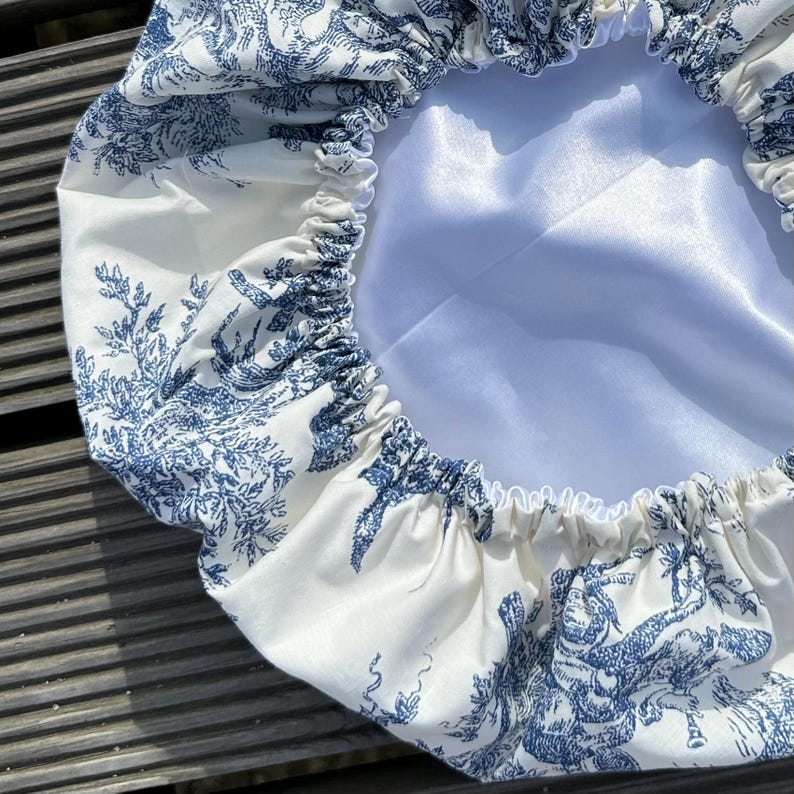 May include: A white and blue toile-patterned bonnet. The bonnet features a blue pastoral scene print on a white background. The interior is a solid white satin material. The edges are gathered with elastic.