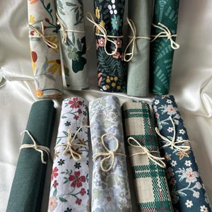 May include: A collection of ten fabric scraps rolled up and tied with twine. The fabrics feature various floral patterns in shades of green, white, and brown.