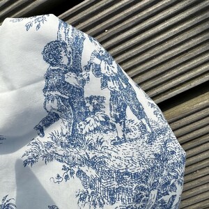 May include: White fabric with a blue toile pattern featuring figures in a pastoral scene. The design includes trees, figures, and foliage, creating a classic, detailed aesthetic. The fabric is folded, showing the pattern's repeat.