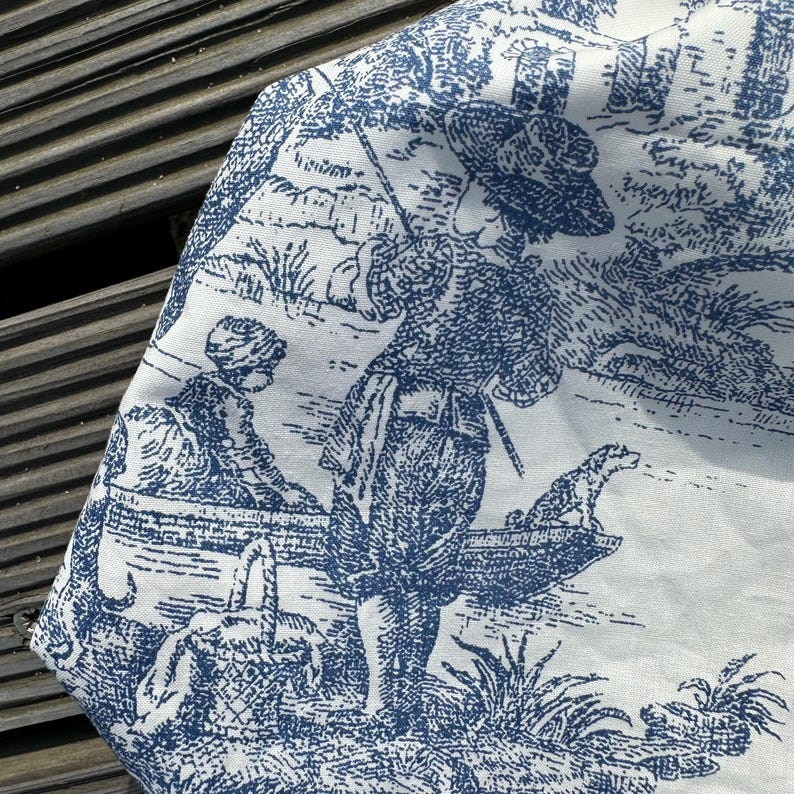 May include: White fabric with a blue toile print depicting a pastoral scene. The design features figures, a dog, and a basket near a body of water. The fabric is folded, showing the detailed pattern and texture.