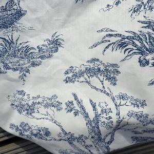 May include: White fabric with a blue toile pattern featuring trees, boats, and landscapes. The design is reminiscent of classic French country scenes, creating a vintage aesthetic. The fabric has a slightly wrinkled texture.