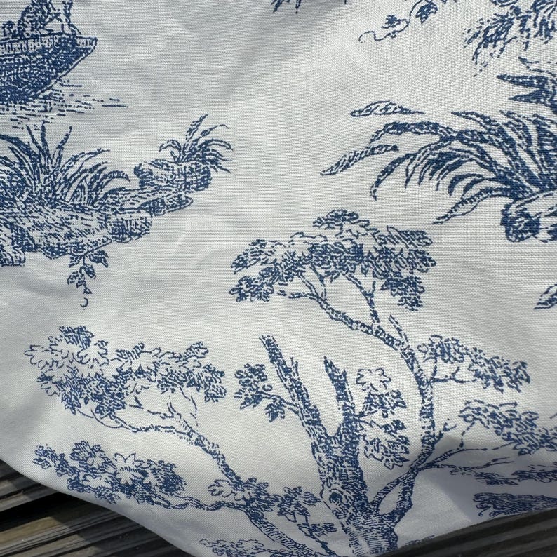 May include: White fabric with a blue toile pattern featuring trees, boats, and landscapes. The design is reminiscent of classic French country scenes, creating a vintage aesthetic. The fabric has a slightly wrinkled texture.