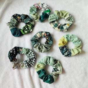 Può includere: Sette elastici per capelli verdi con vari motivi, tra cui floreali, a righe e a quadri. Gli elastici sono realizzati in tessuto nelle tonalità del verde, bianco e giallo. Gli elastici sono disposti su una superficie bianca.