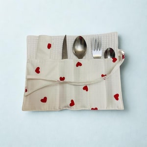 Może przedstawiać: Biały materiałowy uchwyt na sztućce z czerwonymi wzorami serc. Uchwyt zawiera nóż, łyżkę, widelec i małą łyżeczkę. Tkanina ma strukturę wafelka. Uchwyt jest zamykany klapką.