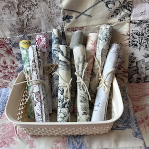 Peut inclure: Un ensemble de rouleaux de tissu dans un panier crème. Les tissus présentent divers motifs, notamment des scènes florales et pastorales, attachés avec de la ficelle. Le panier repose sur un fond de tissu à motifs, créant une ambiance vintage.