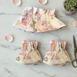 Könnte beinhalten: Gezeigt werden florale Stoffartikel, darunter ein größeres Stück und zwei kleinere. Der Stoff weist ein Muster aus rosa Rosen auf cremefarbenem Hintergrund auf. Verstreut sind rosa Rosenblätter und eine kleine Sukkulentenpflanze in einem grauen Topf.