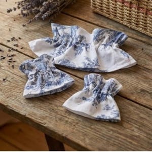 Puede incluir: Tres posavasos de tela toile blanca y azul con bordes con volantes están dispuestos sobre una superficie de madera rústica. Los posavasos presentan un diseño de paisaje clásico azul y blanco. Lavanda seca y una cesta de mimbre en el fondo.