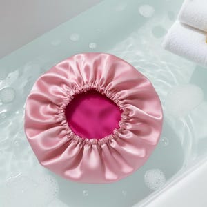 Puede incluir: Un gorro de satén rosa con un interior fucsia en contraste. El gorro es redondo con un borde elástico, diseñado para proteger el cabello. La imagen muestra el gorro flotando en el agua, con burbujas y toallas blancas dobladas en el fondo.