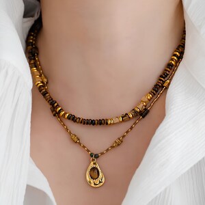 May include: A layered necklace with a gold pendant featuring a brown gemstone. The necklace is made of brown and gold beads.