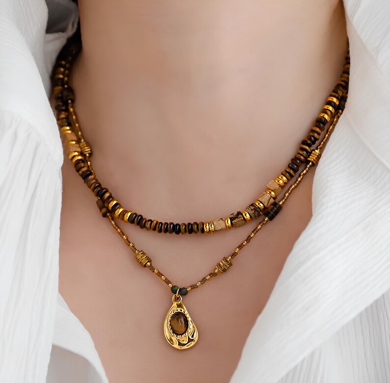 May include: A layered necklace with a gold pendant featuring a brown gemstone. The necklace is made of brown and gold beads.