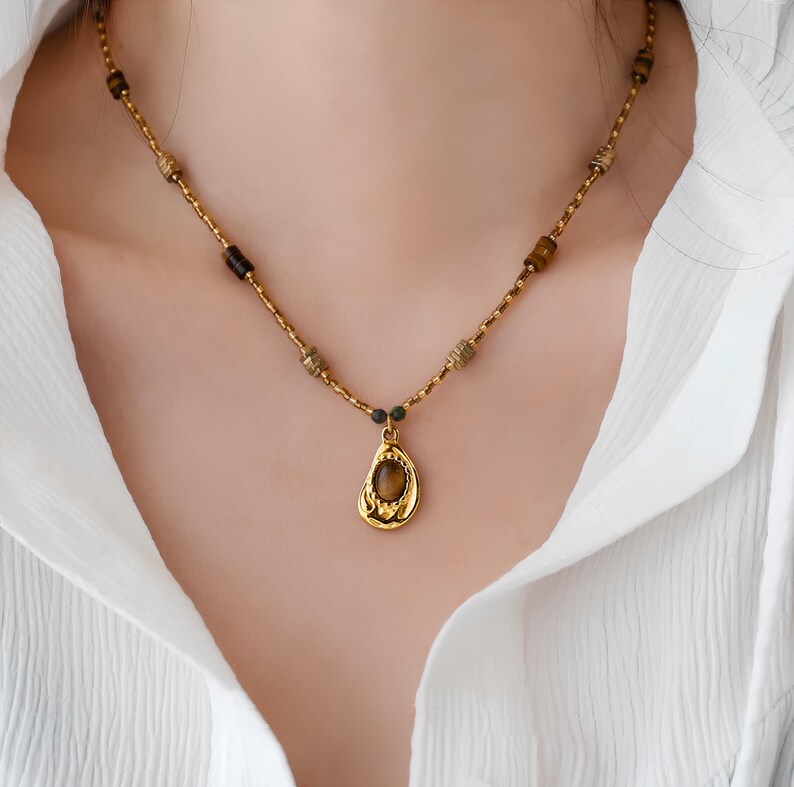 May include: A gold necklace with a teardrop-shaped pendant featuring a brown gemstone. The necklace is made of small gold beads and a chain.