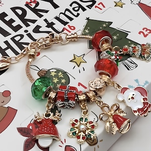 Könnte beinhalten: Goldfarbenes Weihnachts-Charm-Armband mit festlichen Charms. Zu den Charms gehören ein Weihnachtsmann, ein Schneeflocke, eine Glocke, ein Geschenk sowie rote und grüne Perlen. Das Armband wird auf einem Hintergrund mit den Worten "Merry Christmas" angezeigt.
