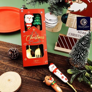 May include: A red Christmas gift set box with gold utensils, a Santa Claus figure, and a Christmas tree. The box reads "Merry Christmas" in gold script. Decorative Christmas-themed serving utensils with snowman and reindeer designs are also visible.