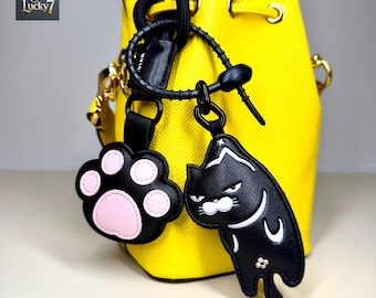 May include: A bright yellow bucket bag with a black strap and gold-colored hardware. Two black leather charms hang from the bag: a paw print with pink pads and a black cat with white details. The Lucky7 logo is in the upper left corner.